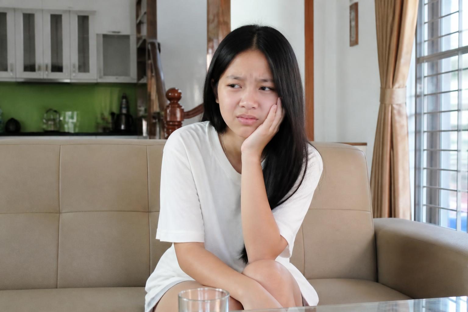 Thiếu nữ ôm má vì đau răng khi ngồi ở nhà, dấu hiệu cần điều trị trước khi niềng răng - ảnh minh hoạ